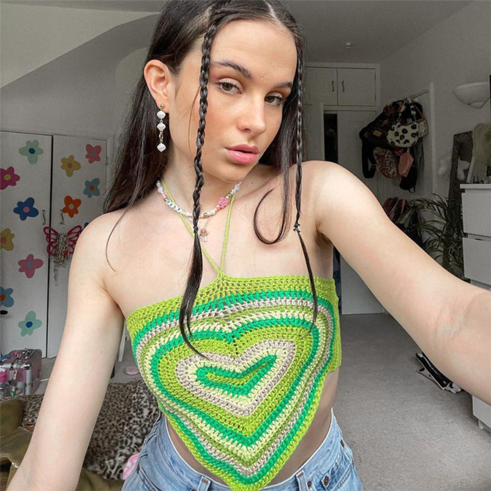 A young woman with long dark braided hair taking a mirror selfie, wearing a vibrant green and white heart-patterned crochet halter top and light-wash denim shorts. She is standing in a brightly decorated room with colorful wall decals and a butterfly-themed background.