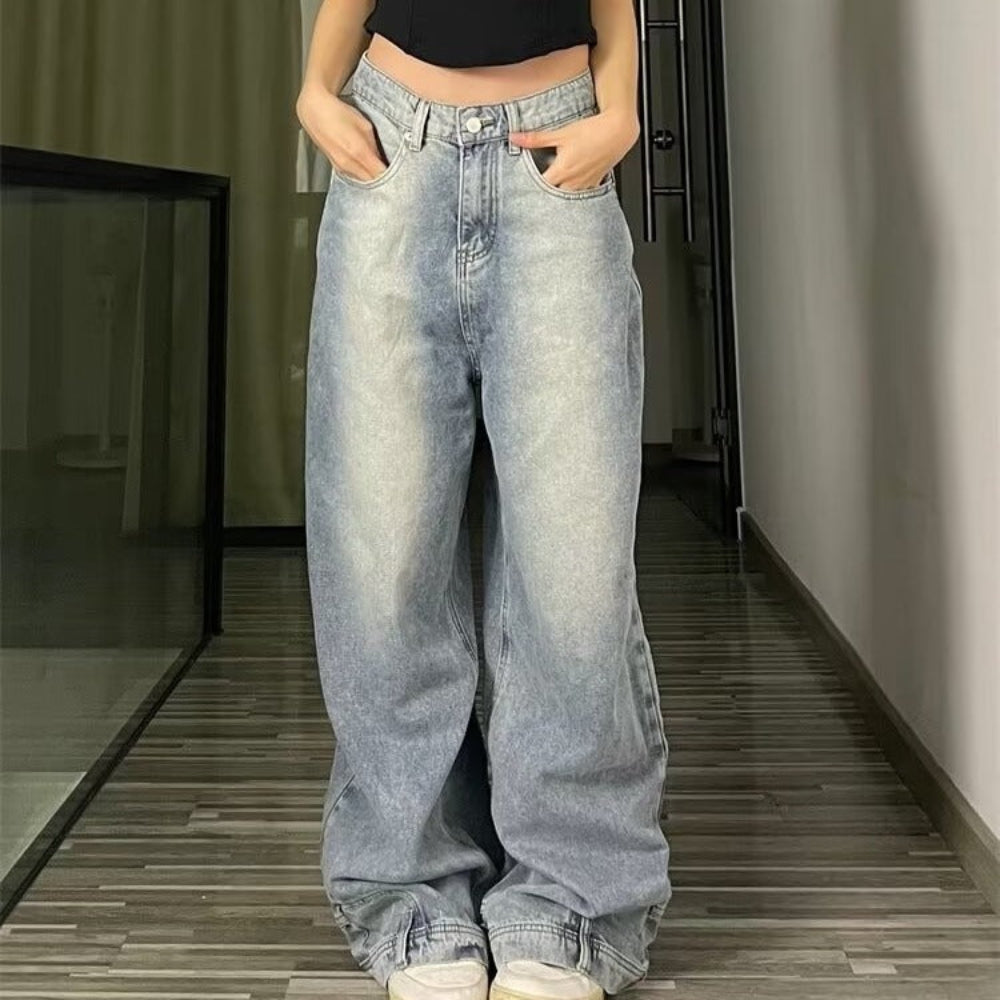 Woman modeling light-wash blue wide-leg baggy jeans with hands in front pockets, paired with a black crop top and white sneakers, standing in a hallway with wooden flooring.