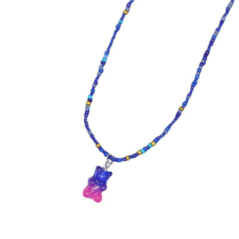 Blue beaded necklace featuring various shades of blue seed beads with gold and turquoise accents, paired with a sparkling translucent blue-to-pink ombre gummy bear pendant. Playful single-strand design laid flat on a white background, showcasing its vibrant and whimsical style.