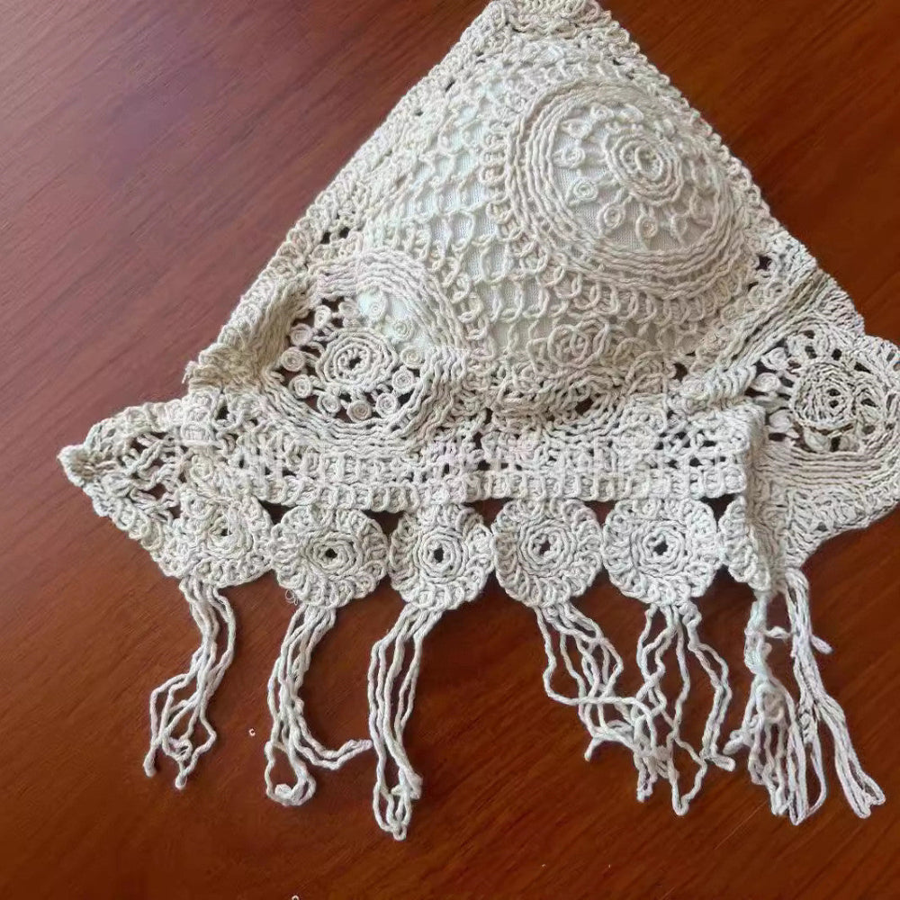 A close-up view of a handmade white crocheted bikini top laid on a wooden surface, showcasing intricate lace patterns, circular motifs, and detailed openwork stitching on the triangle cup. The bottom edge features a decorative band with crocheted circular elements and delicate fringe tassels.
