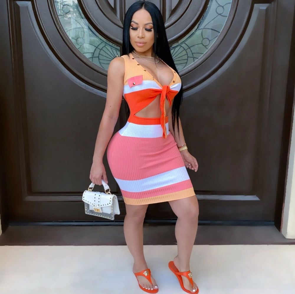 Model wearing a vibrant ribbed knit bodycon mini dress featuring bold horizontal stripes in coral pink, orange, white, and yellow. Sleeveless style with a deep neckline, waist cut-out, and tie accent. Full-body fashion pose holding a small white handbag, styled with orange heels in front of a large brown decorative door.