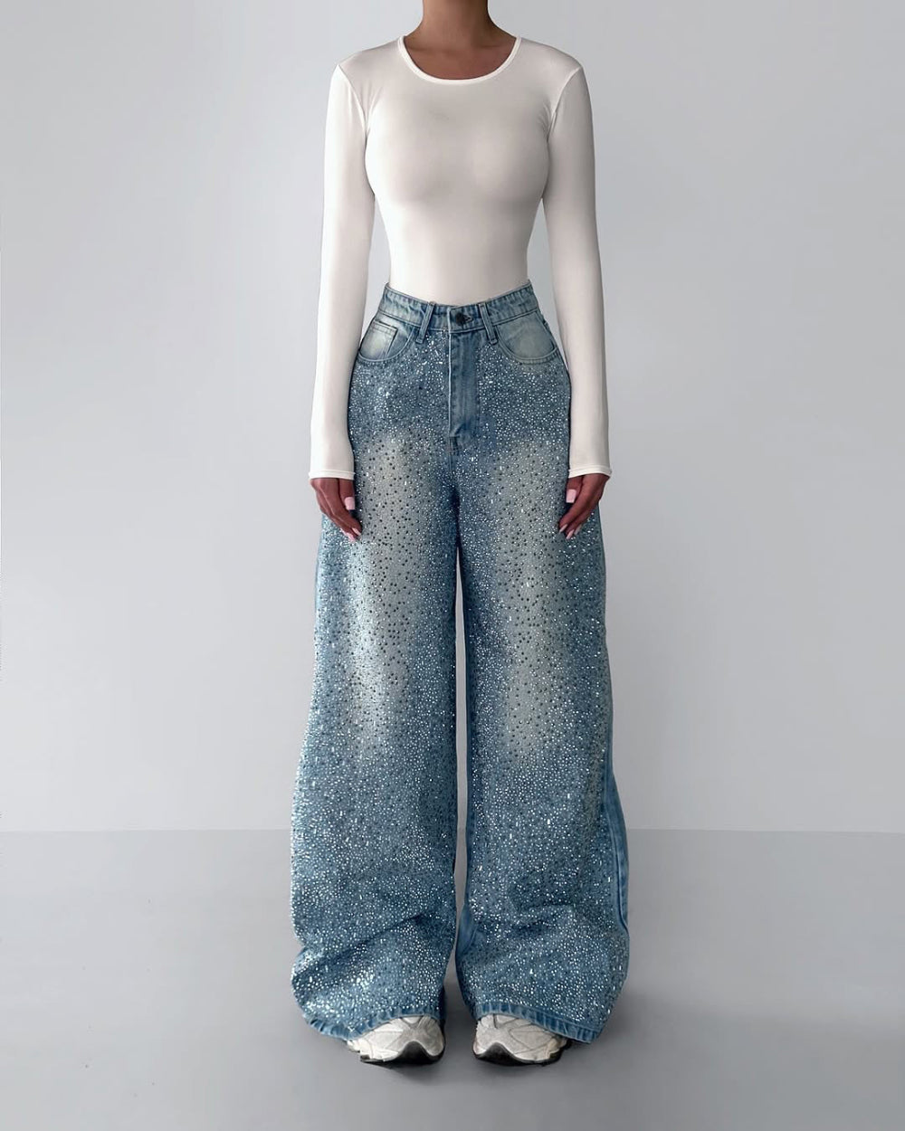 Woman modeling a white long-sleeve bodysuit and light blue wide-leg baggy jeans with acid wash and glitter splatter effect, styled with white sneakers. Full-body minimalist studio fashion shot for Boutique Jolie.