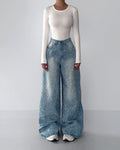 Woman modeling a white long-sleeve bodysuit and light blue wide-leg baggy jeans with acid wash and glitter splatter effect, styled with white sneakers. Full-body minimalist studio fashion shot for Boutique Jolie.