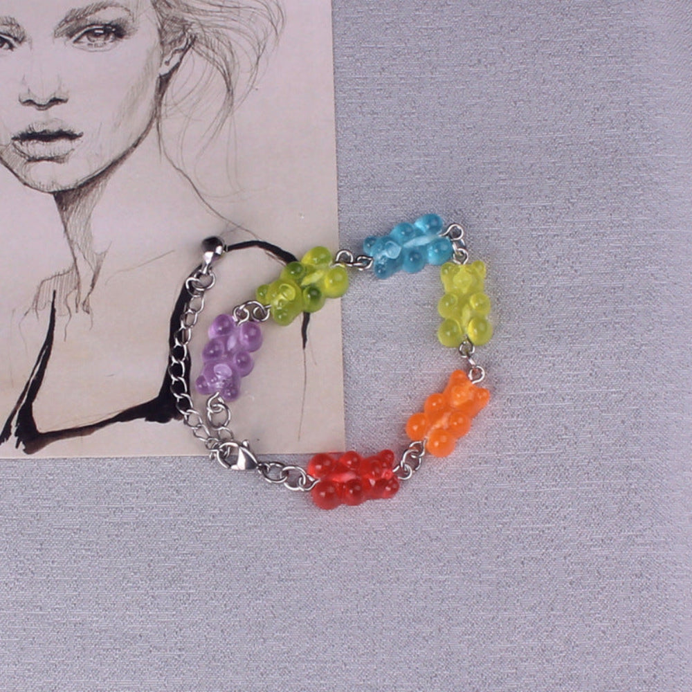 Rainbow gummy bear charm bracelet with silver metal chain, lobster clasp, and black tassel, featuring translucent colorful gummy bear charms arranged in rainbow order (purple, green, blue, yellow, orange, red), displayed on a textured background.