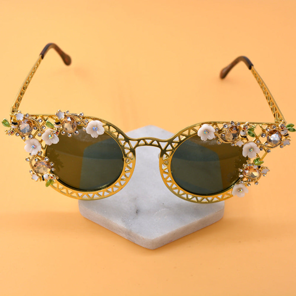 Elegant gold metal cat-eye sunglasses featuring intricate lattice frames, white flowers, green leaves, sparkling crystals, and dark green tinted lenses, displayed on marble