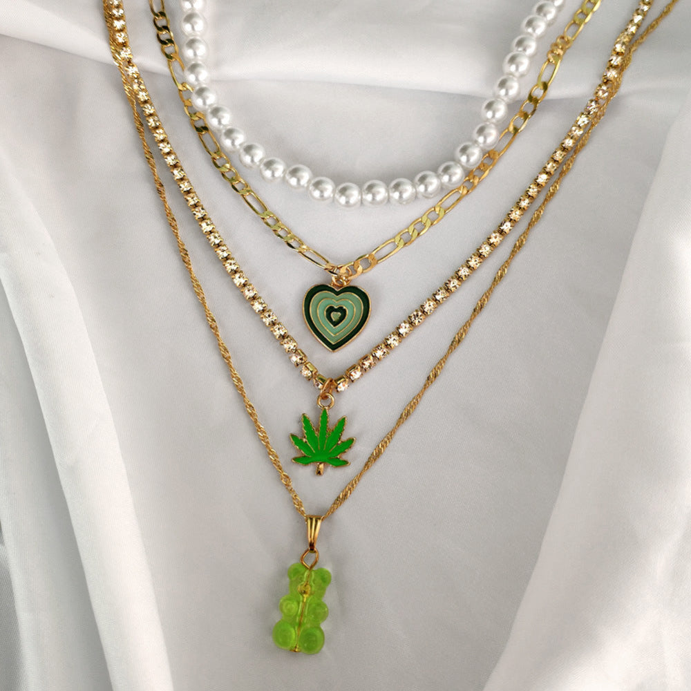 Gold pearl layered necklace stack with green heart, leaf & gummy bear pendants – trendy mixed metal statement jewelry on white background.
