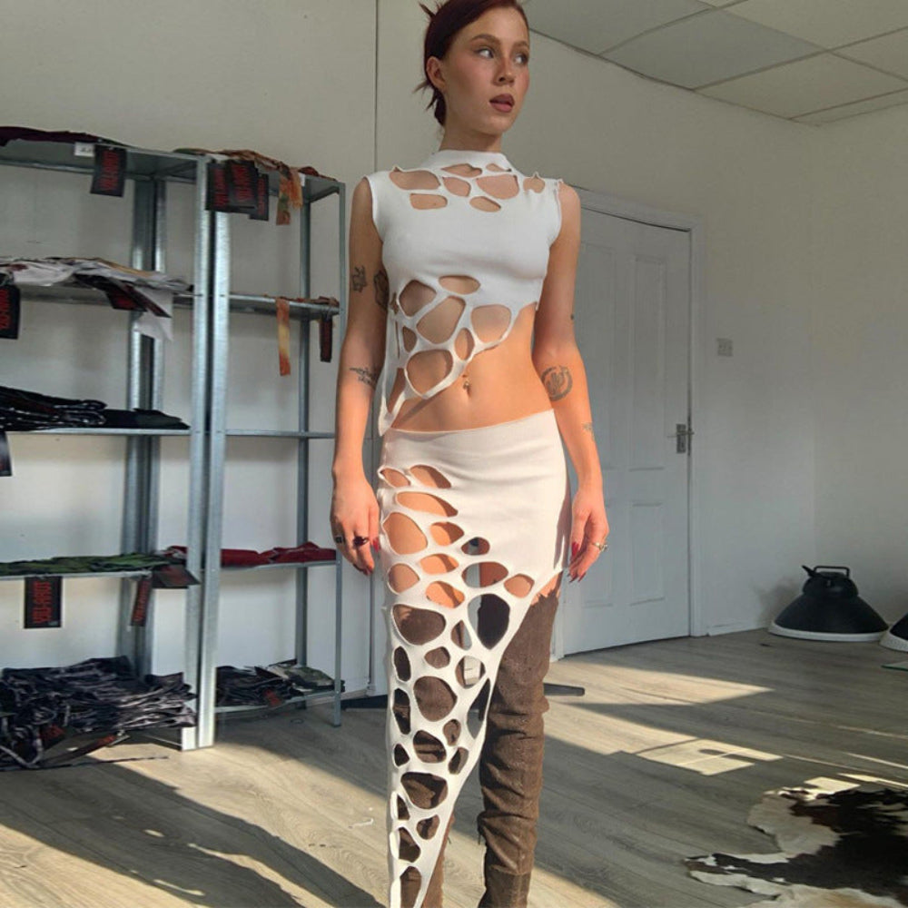 Model wearing a bold white two-piece outfit with artistic perforated cut-out designs across the sleeveless crop top and matching skirt. Full-body pose in a bright workspace with metal shelves and natural light, styled with brown over-the-knee boots.