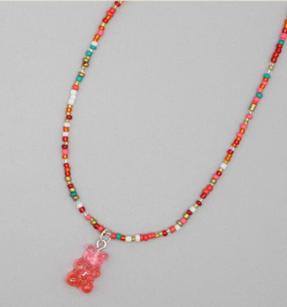 Colorful beaded necklace featuring a mix of vibrant pink, red, orange, white, teal, and gold seed beads with a translucent pink gummy bear pendant. The playful single-strand necklace is displayed flat on a light gray background, highlighting its fun and whimsical design.