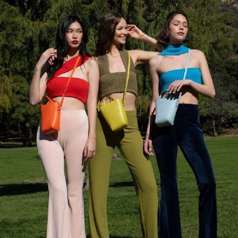 Three models posing outdoors wearing ribbed knit crop tops in red one-shoulder, olive green sleeveless with front cutout, and blue strapless turtleneck styles, paired with color-coordinated flared pants and matching crossbody bags.