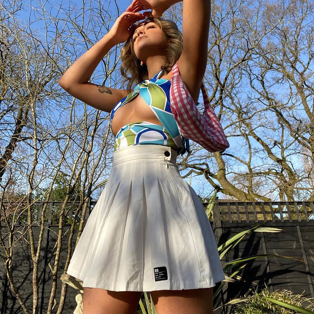 Woman with blonde hair posing outdoors on a sunny day, wearing a colorful geometric print halter crop top with cut-out detail and a white pleated mini skirt. She is adjusting sunglasses on her head while carrying a pink gingham crossbody bag, with bare trees and a blue sky in the background