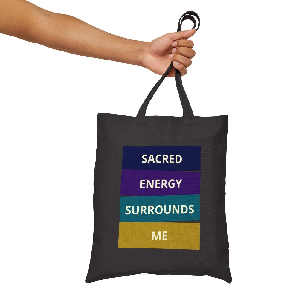 Black canvas tote bag with "SACRED ENERGY SURROUNDS ME" spiritual quote in colorful stacked blocks (navy, purple, teal, gold), held by hand against white background.