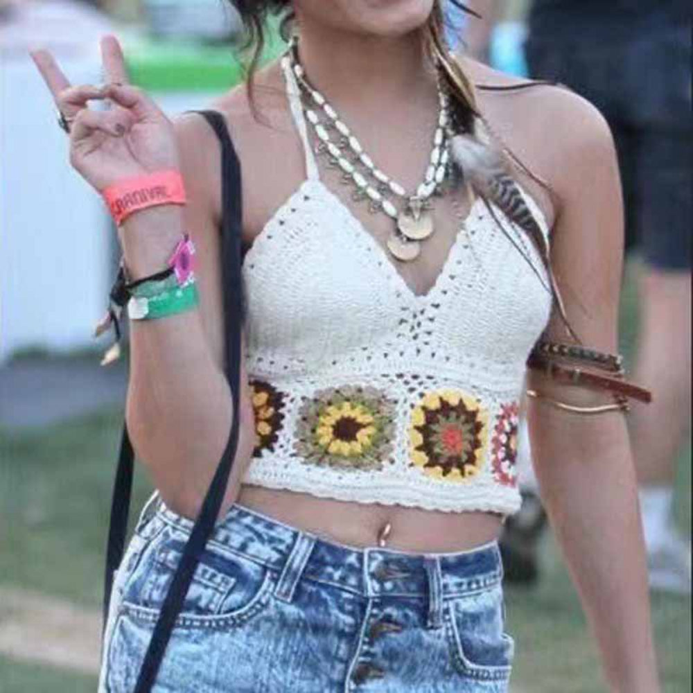 A woman wearing a boho white crochet halter crop top with intricate openwork stitching and colorful floral granny square motifs along the hem. Styled with light-wash denim jeans and layered festival jewelry, she poses making a peace sign, capturing a fun, summery, free-spirited vibe.