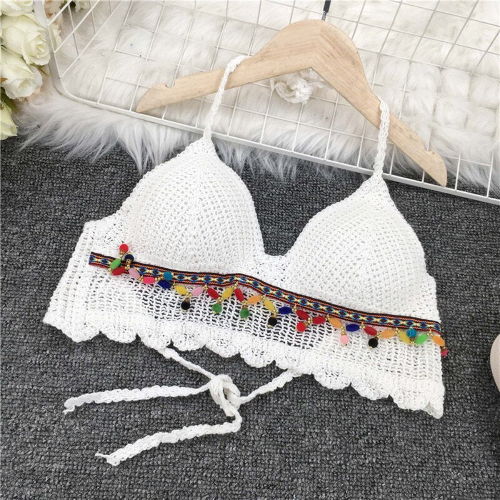 White crochet halter crop top with colorful pom pom trim and tie front hanging on wooden hanger.