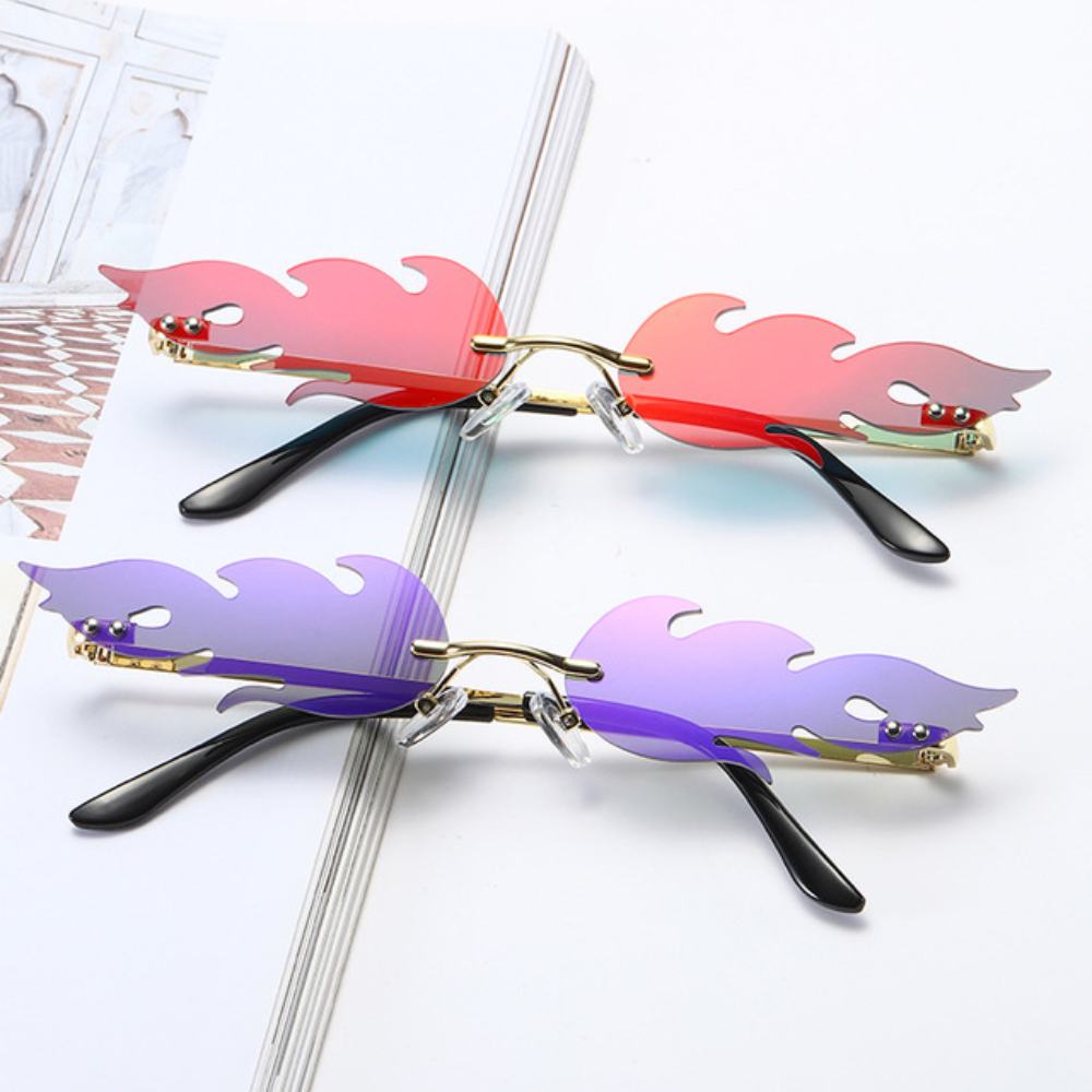 Two pairs of rimless flame sunglasses in red and purple with gold temples, styled on an open magazine.