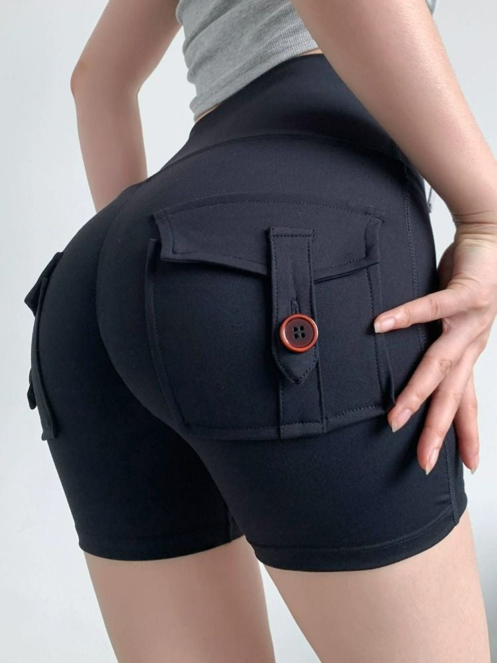 Woman modeling black high-waisted cargo stretch shorts with back flap pockets and button detail, shown from a rear three-quarter view with hand on hip against a clean white studio background.