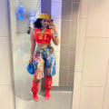 Woman with curly black hair taking a mirror selfie in a modern hallway, wearing a yellow cap, red Chicago 12 crop top, colorful paint-splatter cargo shorts, red thigh-high boots, and holding a blue furry purse. Bold street-style fashion look for Boutique Jolie.