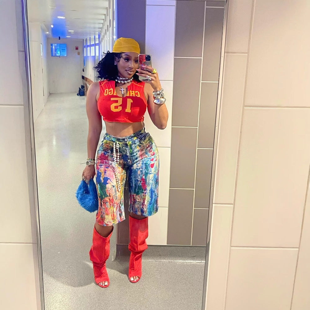 Woman with curly black hair taking a mirror selfie in a modern hallway, wearing a yellow cap, red Chicago 12 crop top, colorful paint-splatter cargo shorts, red thigh-high boots, and holding a blue furry purse. Bold street-style fashion look for Boutique Jolie.