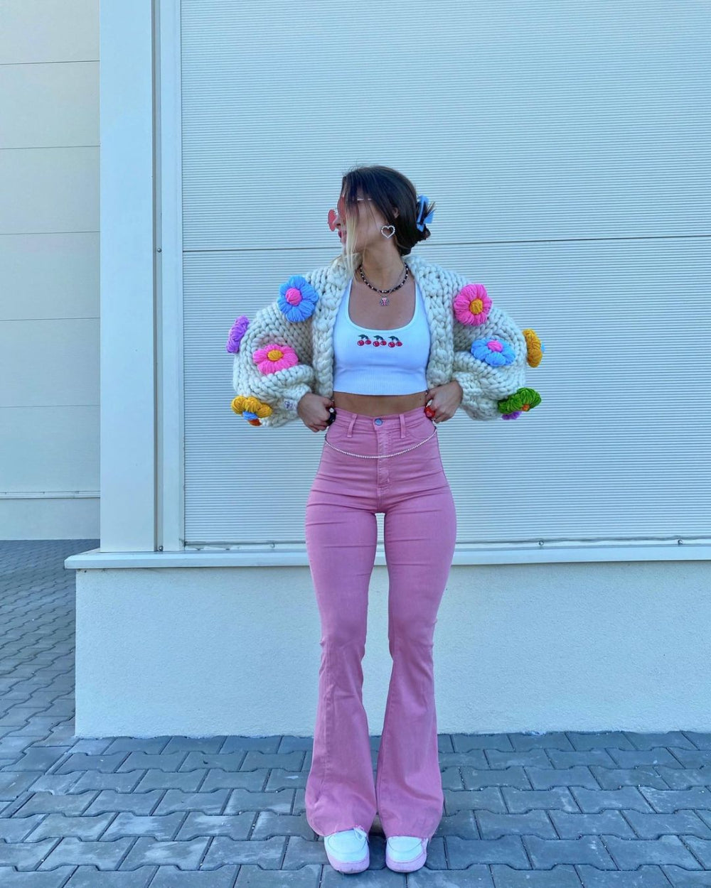 Woman with dark hair posing confidently in a white knit cardigan with colorful pom-poms, white crop top, pink pants, and white sneakers, standing in front of a white slatted wall.