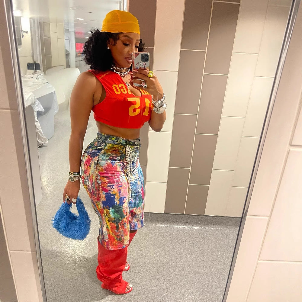 Woman with curly black hair taking a mirror selfie, wearing a yellow cap, red Chicago 12 crop top, multicolored paint-splatter denim shorts, red thigh-high boots, and holding a bright blue furry purse. Stylish streetwear outfit for Boutique Jolie.