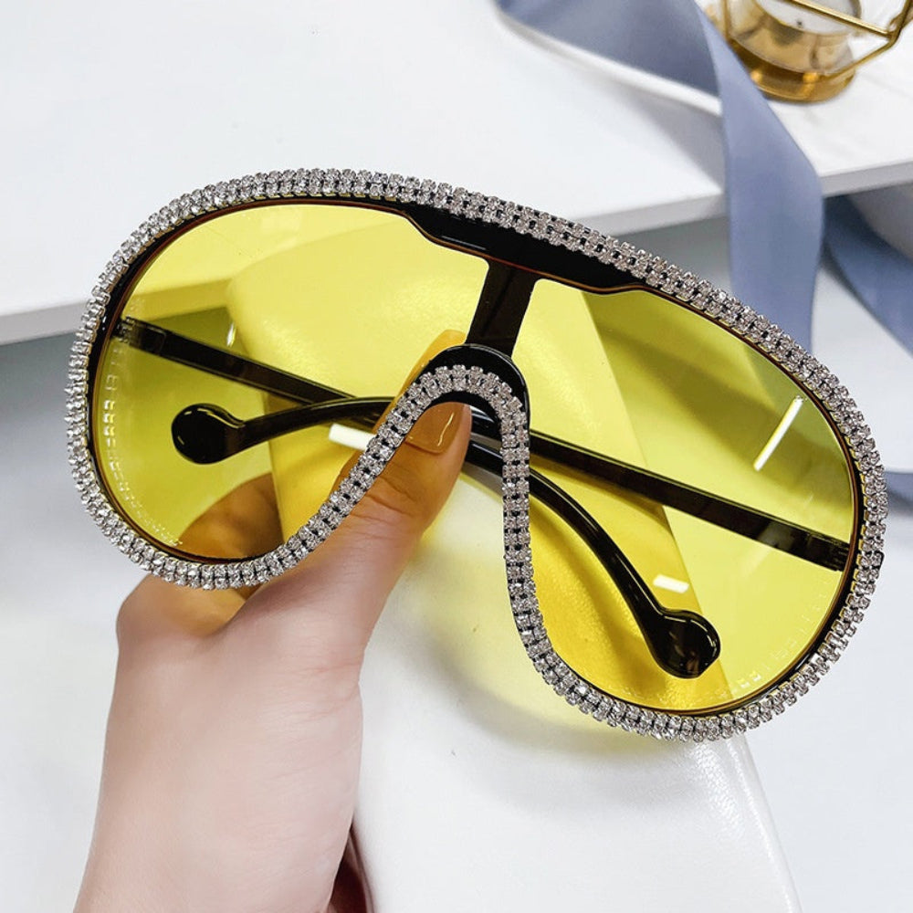 Hand holding oversized yellow-lens shield sunglasses with crystal rhinestone trim.