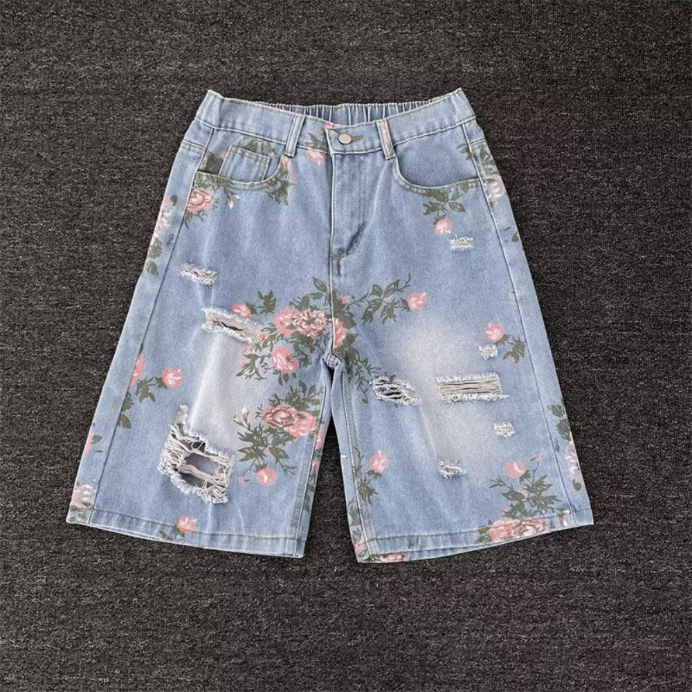 Light blue distressed denim shorts with pink floral rose prints, frayed rips, and raw edges, laid flat on a dark surface. Detailed product view highlighting the vintage-inspired design for Boutique Jolie.