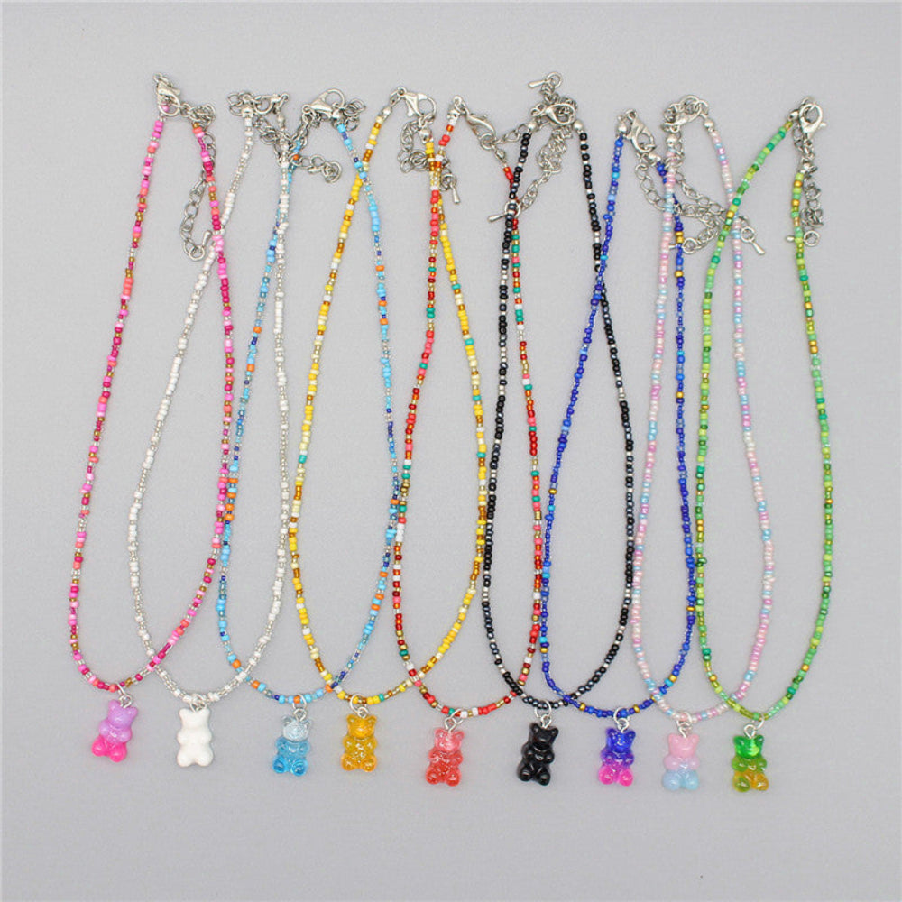 Collection of colorful beaded gummy bear necklaces in various styles — each with vibrant seed beads in matching color themes and a translucent gummy bear pendant. Includes pink, white, blue, yellow, red, black, purple, pastel, and green variations. Silver clasp and extender chain, displayed together on a light gray background.