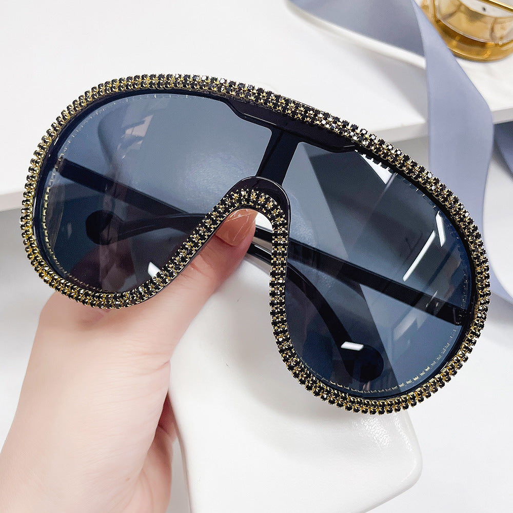 Hand holding oversized dark-lens shield sunglasses with gold rhinestone trim.