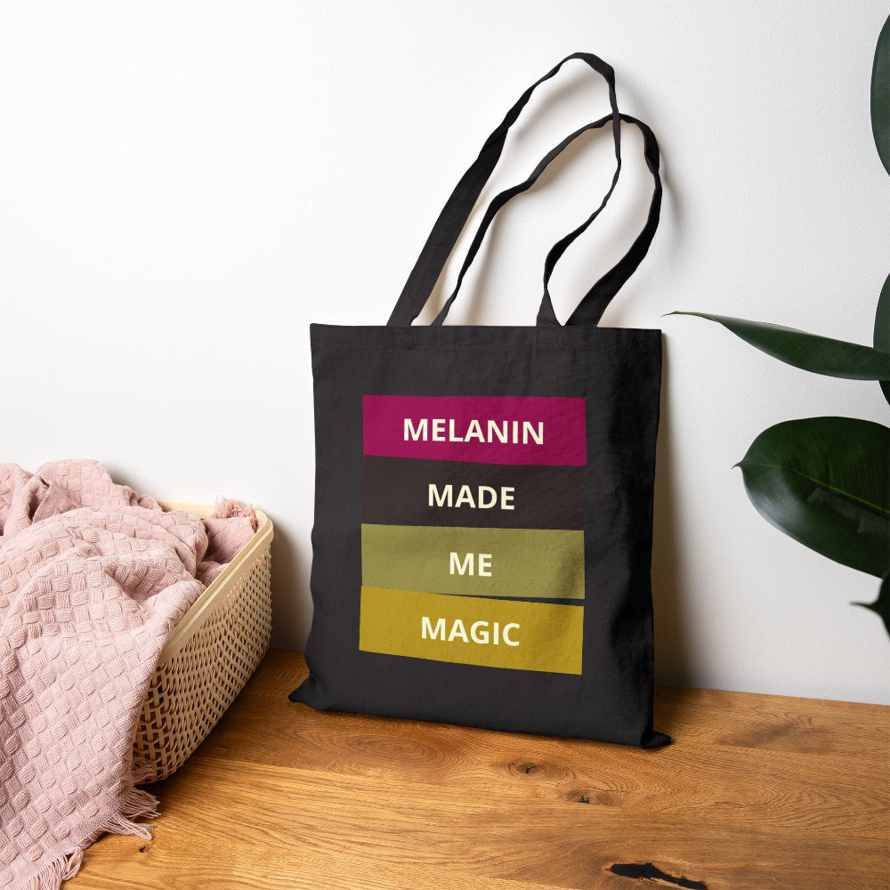 Black canvas tote bag with "MELANIN MADE ME MAGIC" graphic on wooden table with pink throw and plant.