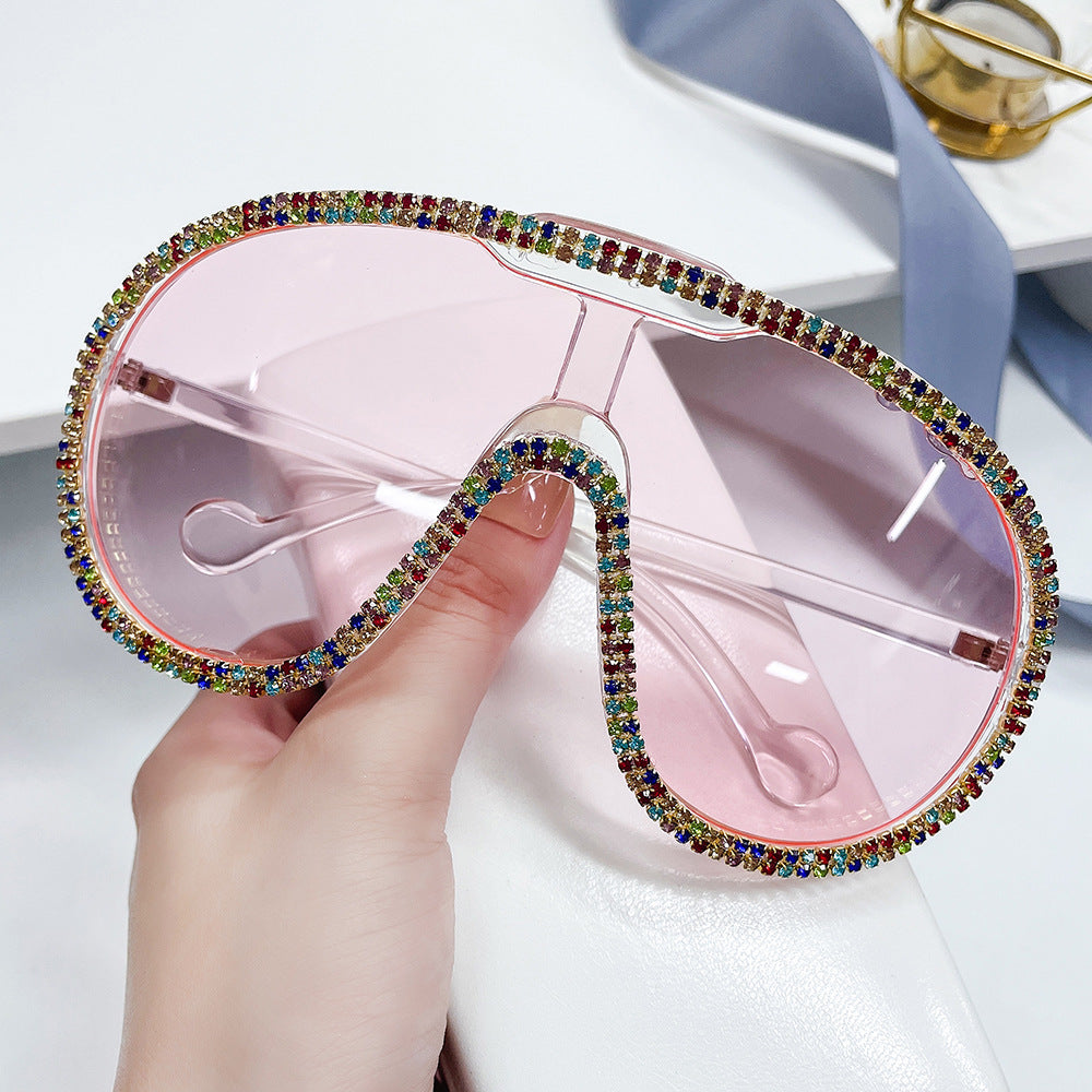 Hand holding oversized pink-lens shield sunglasses with rainbow rhinestone trim.