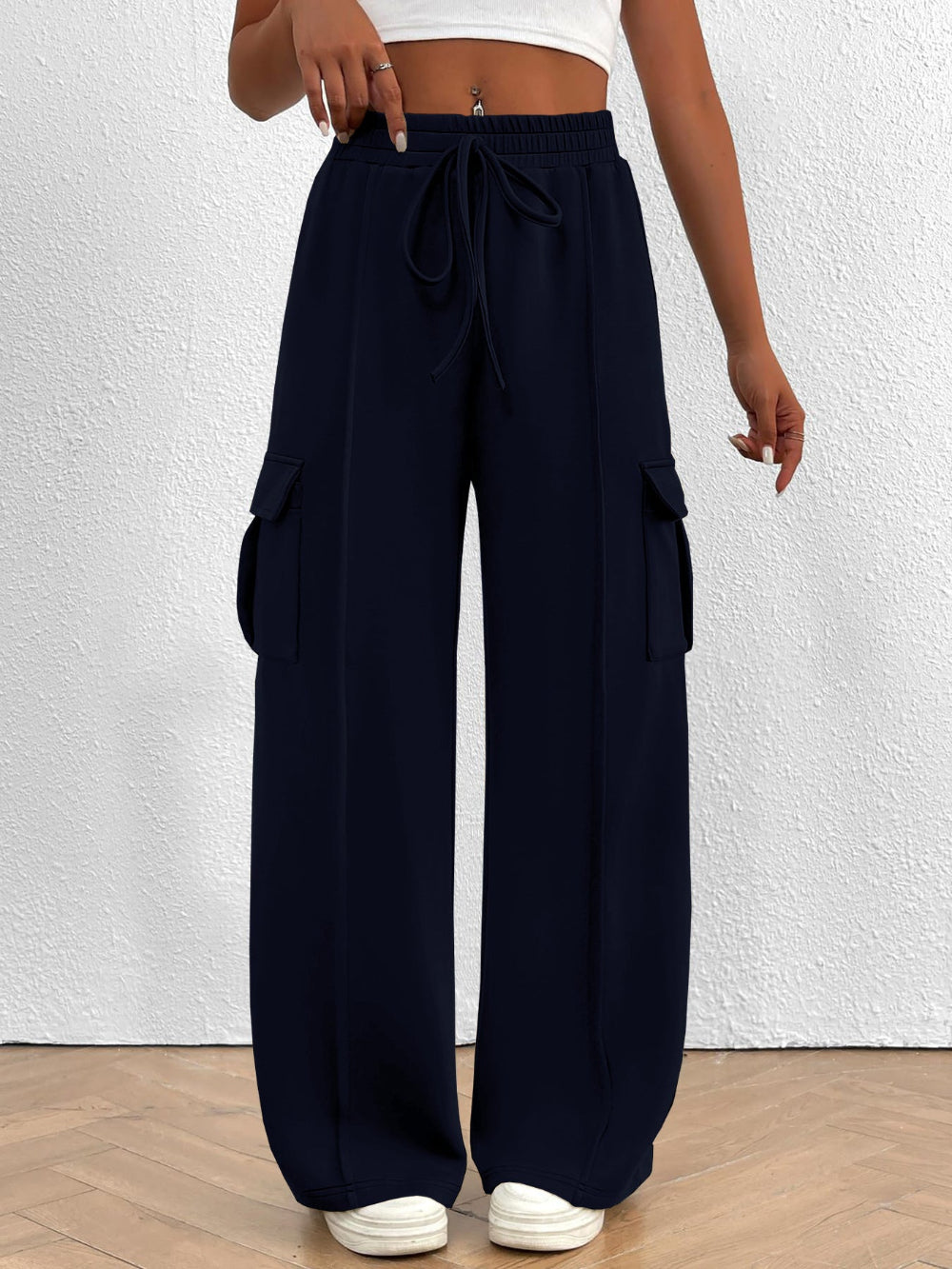 Woman wearing navy blue wide leg cargo pants with drawstring waist and large side pockets paired with a white top and white sneakers against a textured white wall background
