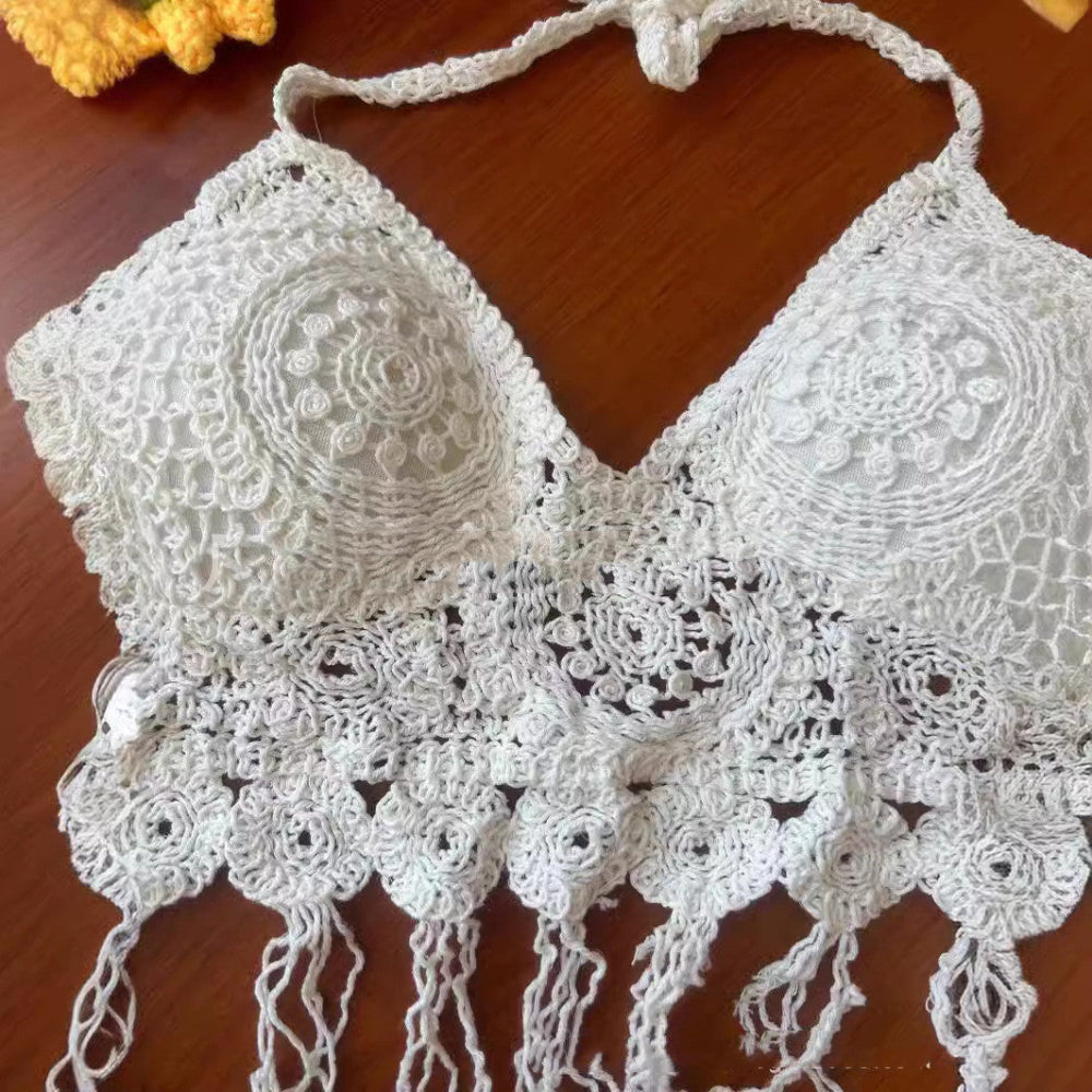 A close-up of a handmade white crocheted bikini top featuring intricate circular lace motifs and detailed openwork patterns on the triangle cups. The top has delicate halter ties and a decorative fringed hem with multiple tassels, beautifully displayed on a wooden surface. A small portion of a yellow crocheted item is visible in the top corner.