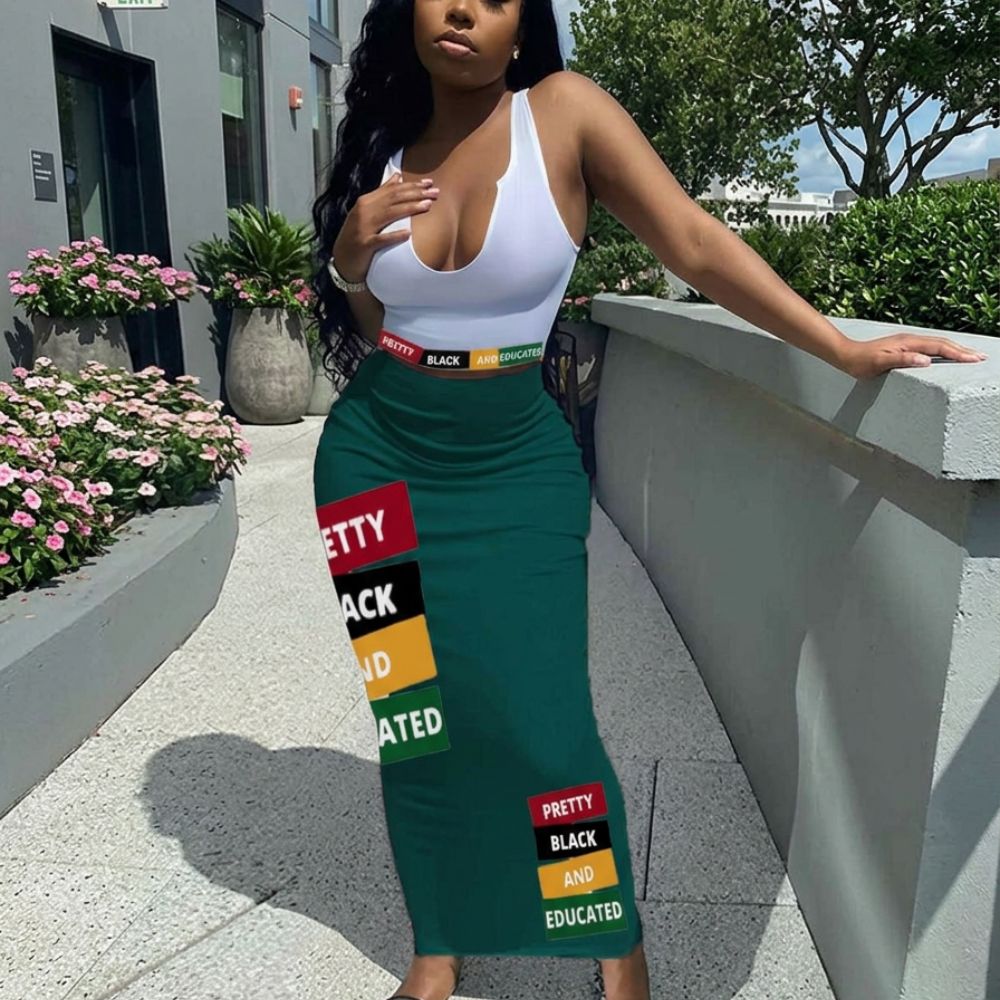 Model in a stylish outfit featuring a fitted white top and high-waisted dark green maxi skirt decorated with bold multicolored rectangular patches reading "PRETTY", "BLACK", and "EDUCATED". Full-body fashion pose outdoors in natural daylight with modern architecture and greenery in the background.