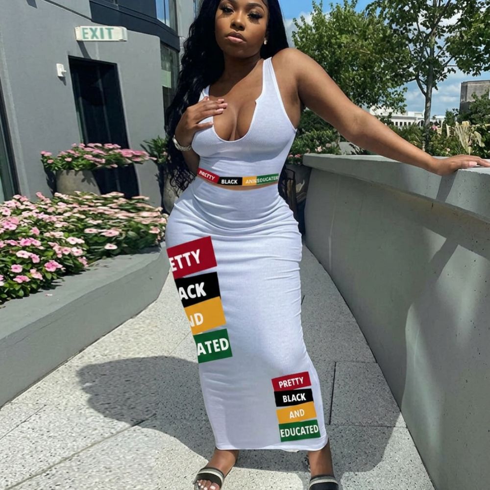 Model in a stylish all-white outfit featuring a deep V-neck sleeveless top and long form-fitting skirt decorated with bold multicolored rectangular patches reading "PRETTY", "BLACK", and "EDUCATED". Full-body fashion pose outdoors in natural daylight with modern architecture and greenery in the background.