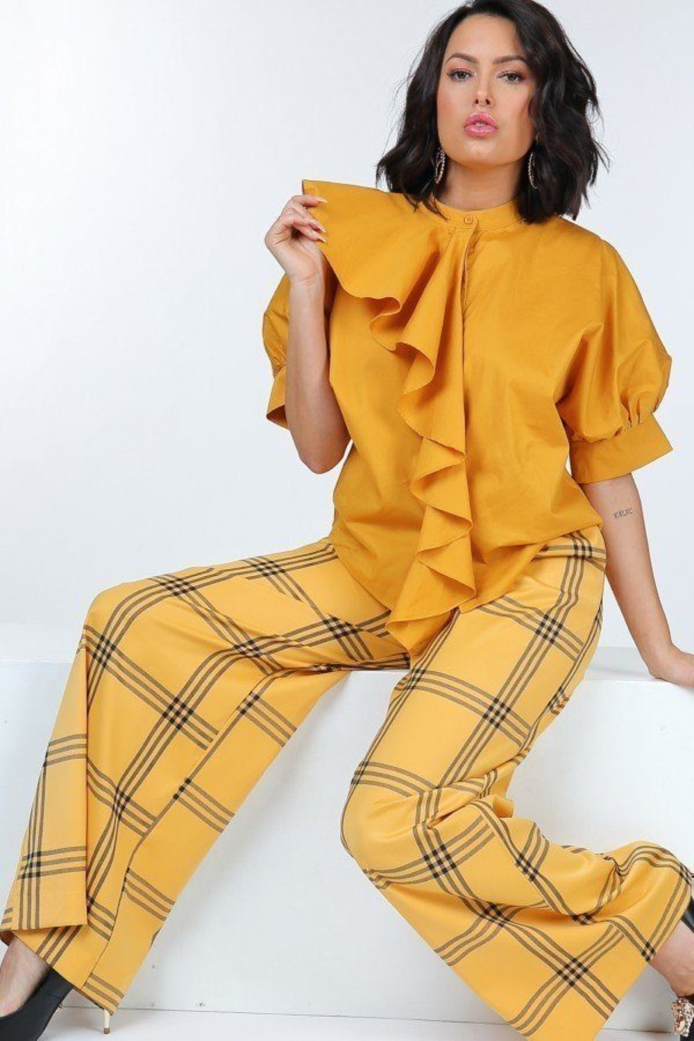 Woman wearing a mustard ruffle blouse with puff sleeves and yellow windowpane plaid wide-leg pants, seated with one hand lifting the ruffle.