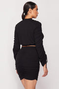 Back view of a woman wearing a black ruched two-piece set with a long-sleeve crop top, drawstring sleeves, and a matching mini skirt.