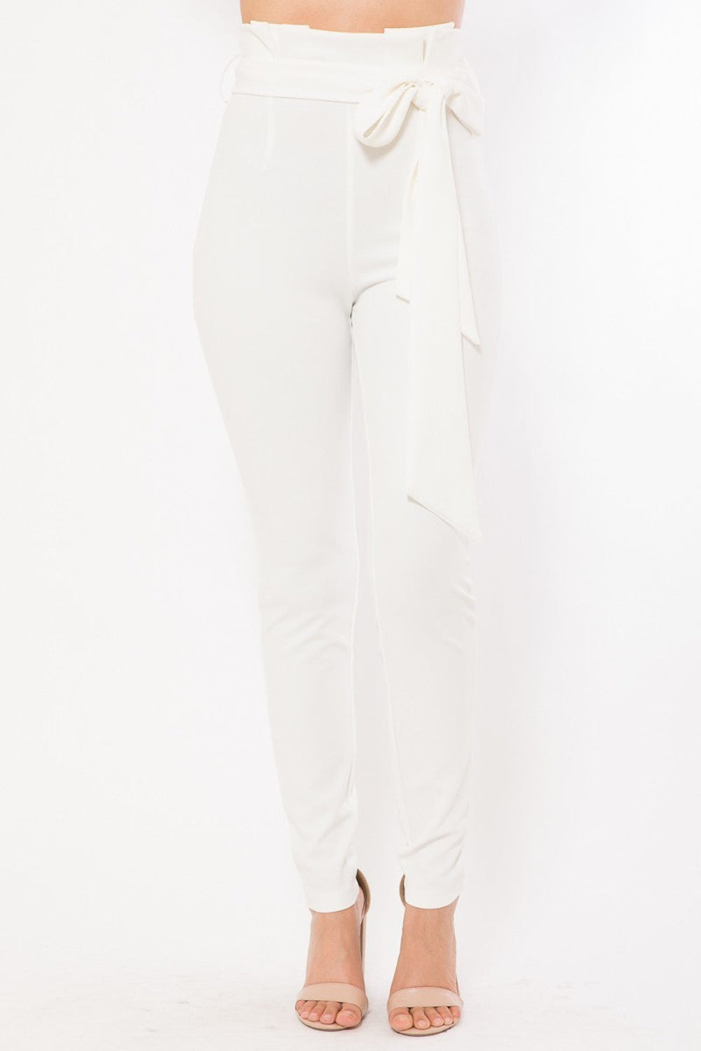 Front view of white high-waist skinny pants with a paperbag waist, side tie belt, and a slim ankle-length fit, worn with nude heels.
