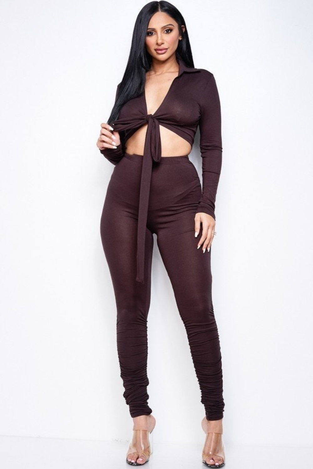 Woman wearing a dark brown two-piece set with a long-sleeve tie-front crop top, ruched skinny pants, and clear heels.