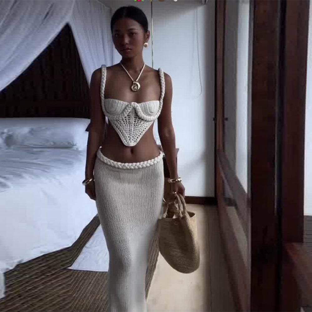 "Model wearing a white crochet knit two-piece set featuring a square-neck crop camisole top with thick straps and a high-waisted ribbed maxi skirt, holding a woven bag in a bedroom setting."
