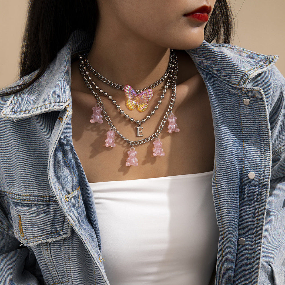 Layered butterfly and pink teddy bear charm necklaces on model in denim jacket – trendy statement jewelry stack, fashion close-up.