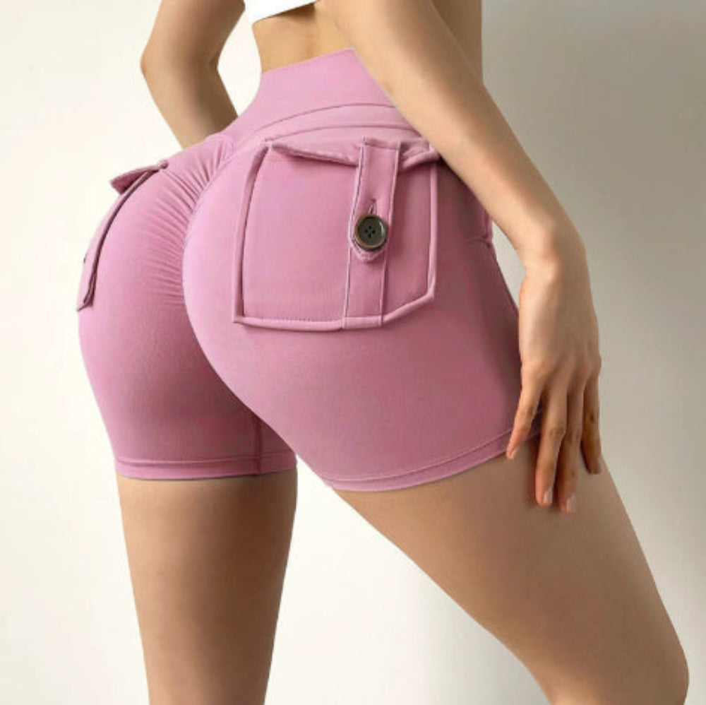 Woman modeling pink high-waisted cargo stretch shorts with a large back flap pocket and button detail, shown from a rear three-quarter view against a plain studio background.
