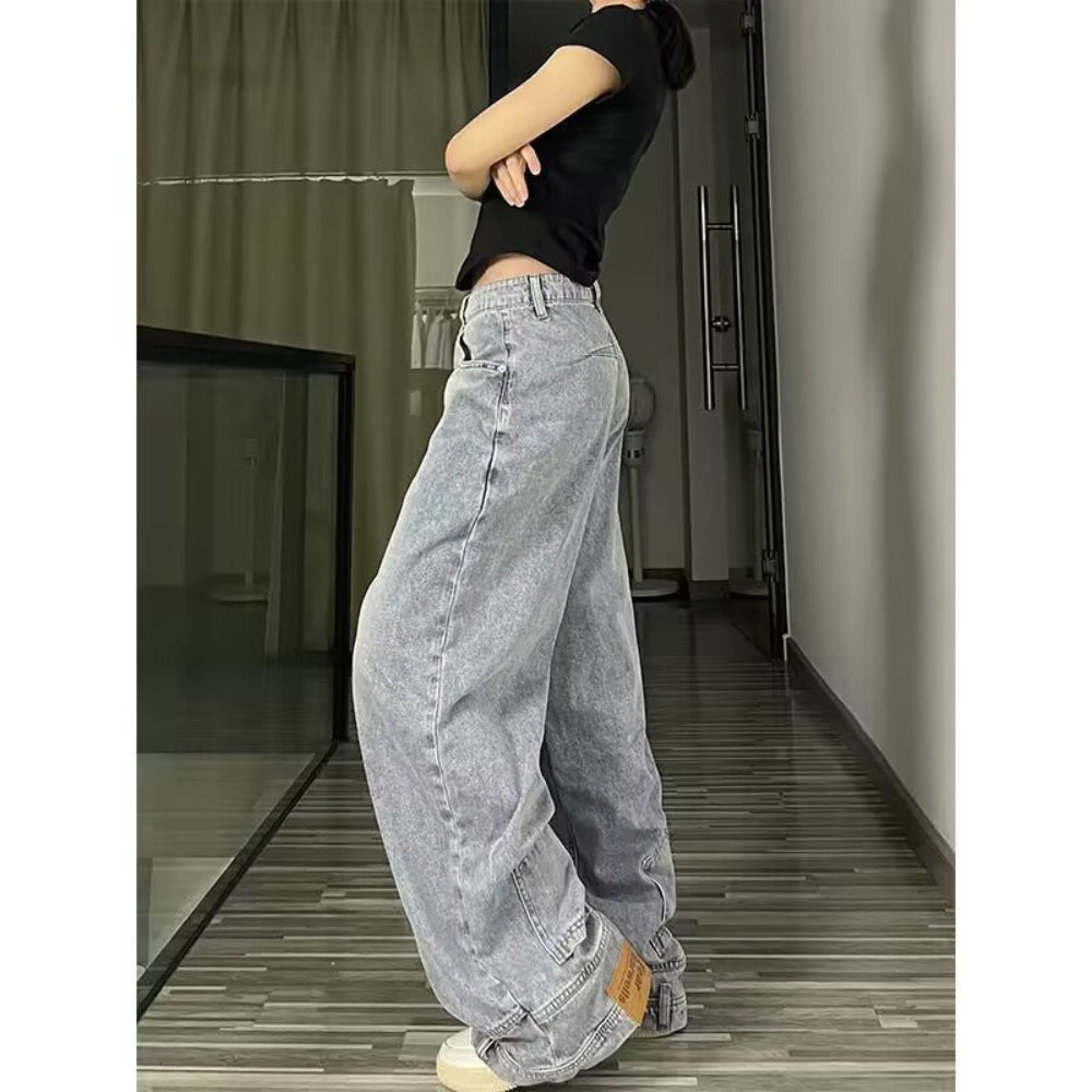Woman modeling light-wash blue wide-leg baggy jeans from a side-back view, paired with a black crop top, standing in an indoor hallway with wooden flooring.