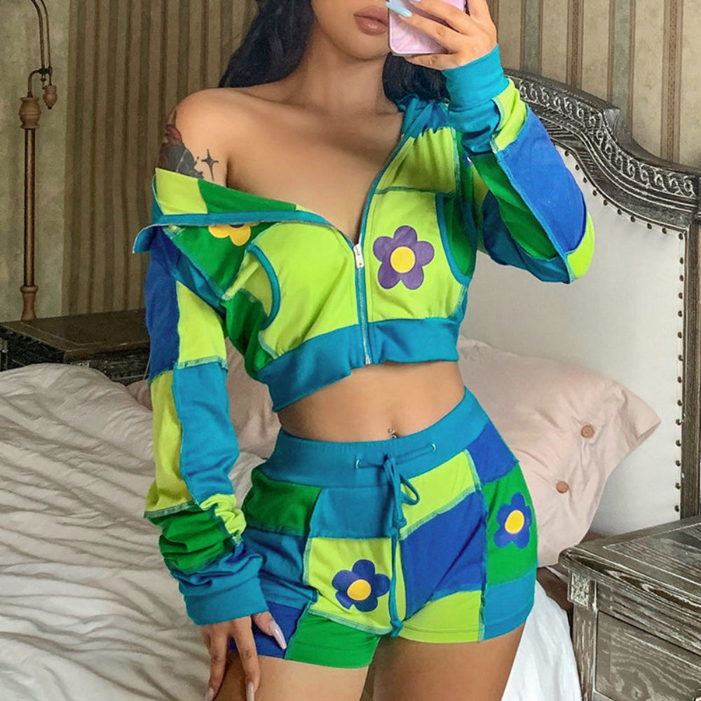 Woman taking a mirror selfie in a vibrant patchwork cropped zip-up hoodie and matching shorts set in lime green, blue, and green panels. The off-shoulder hoodie features colorful daisy flower appliqués, a partial zipper opening, and drawstring hood, while the shorts have a drawstring waist and flower details. Styled with a visible belly button piercing in a bedroom setting.
