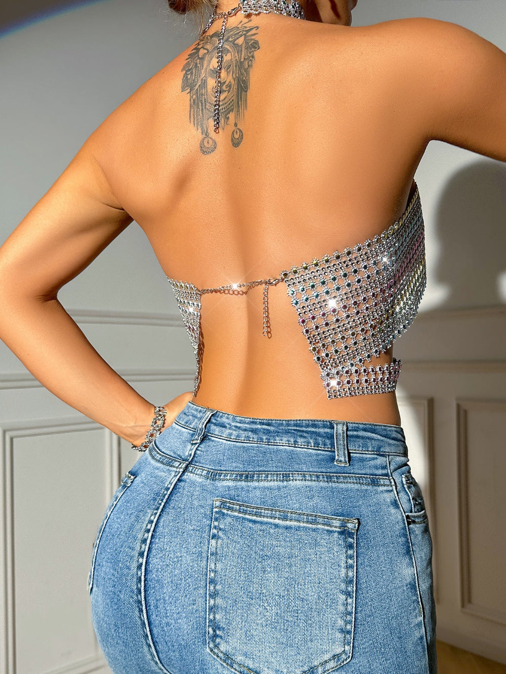 Back view of a woman wearing a sparkling silver rhinestone halter crop top with open back design, chain details, and dangling embellishments. Features a large back tattoo, styled with light blue high-waisted jeans.