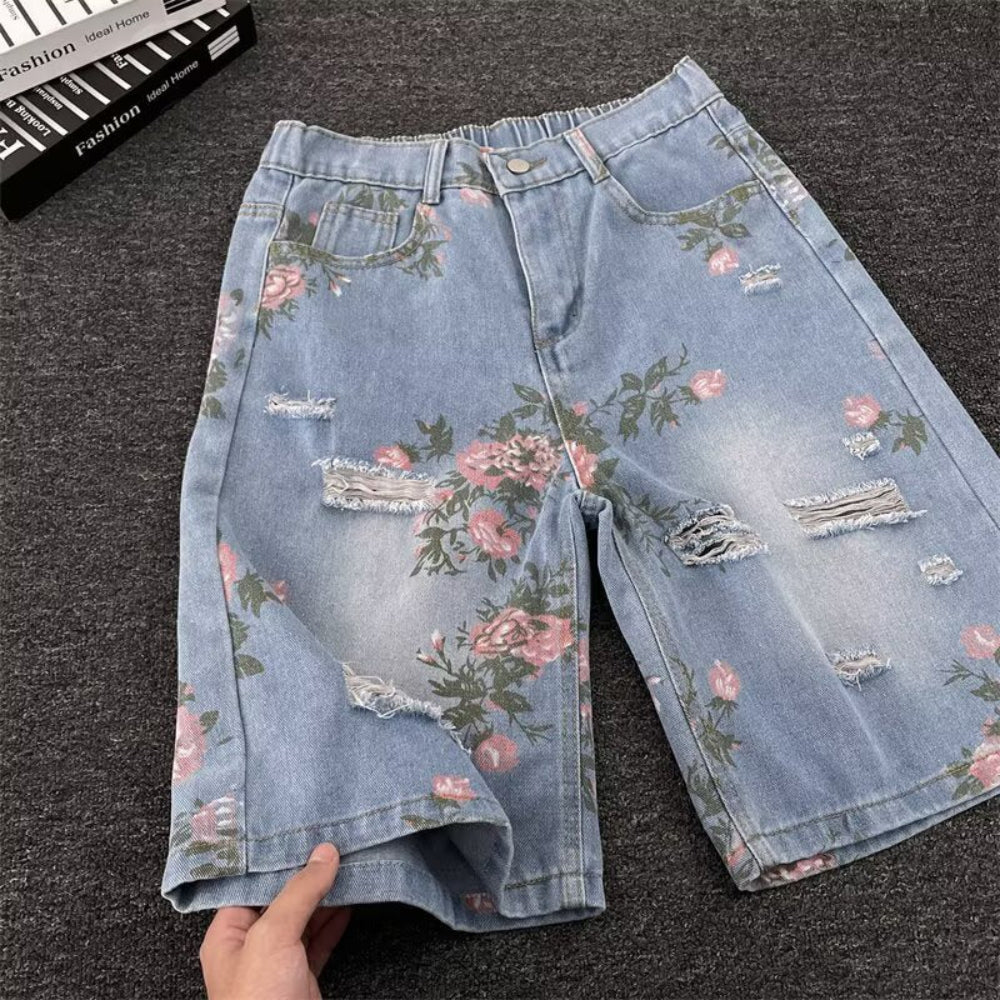 Light blue distressed denim shorts with pink floral rose print and frayed rips, laid flat on a dark surface. Product photography highlighting the vintage-style design and details for Boutique Jolie.