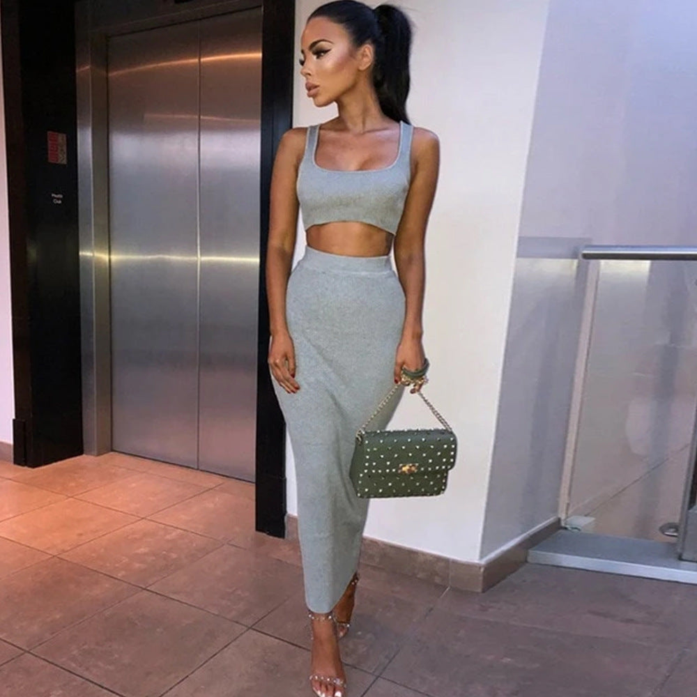 Model in a sleek gray two-piece outfit featuring a sleeveless crop top and form-fitting midi skirt. Full-body side pose with one hand near her face, accessorized with a green handbag and strappy heels, standing in a contemporary building hallway with elevator doors visible.