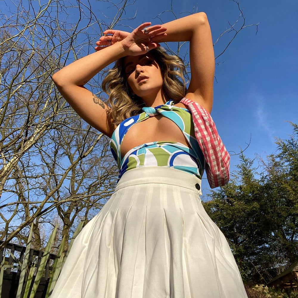 Blue green geometric print halter crop top with front twist knot and plunge neckline, paired with a white pleated skirt, worn by a model posing outdoors.