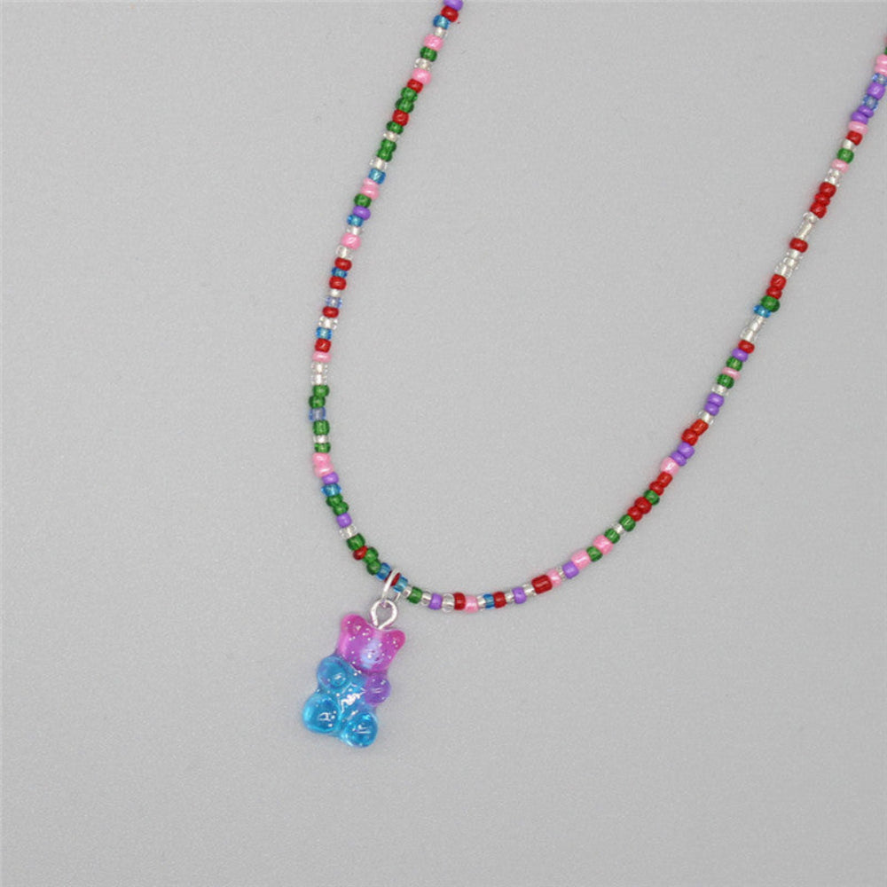 Multicolored beaded necklace with a vibrant mix of red, green, pink, blue, purple, and silver seed beads. Features a translucent two-tone gummy bear pendant in purple and blue. The playful single-strand necklace is displayed flat on a light gray background, showcasing its fun and colorful design.