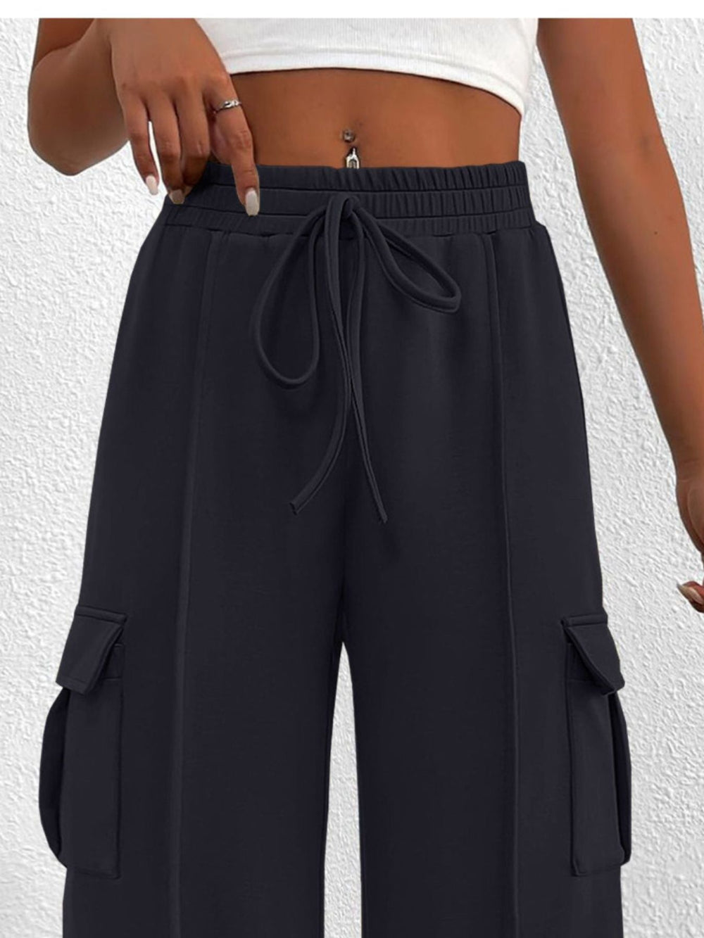 Woman wearing black wide leg cargo pants with drawstring waist and large side pockets against a textured white background