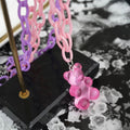 Close-up of a translucent hot pink gummy bear pendant hanging from a pastel pink and purple chunky chain necklace. Displayed on a modern black stand with a stylish black-and-white floral background, highlighting the glossy, playful charm and vibrant chain details.