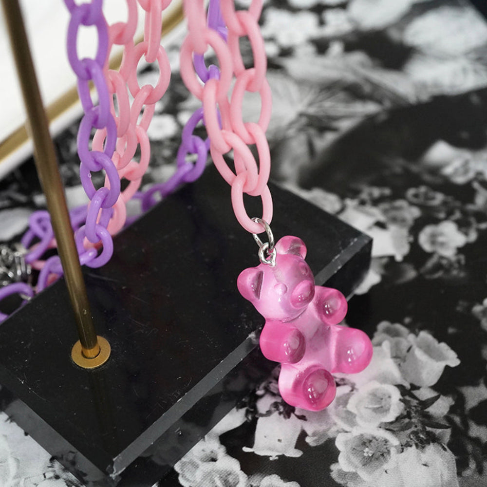 Close-up of a translucent hot pink gummy bear pendant hanging from a pastel pink and purple chunky chain necklace. Displayed on a modern black stand with a stylish black-and-white floral background, highlighting the glossy, playful charm and vibrant chain details.
